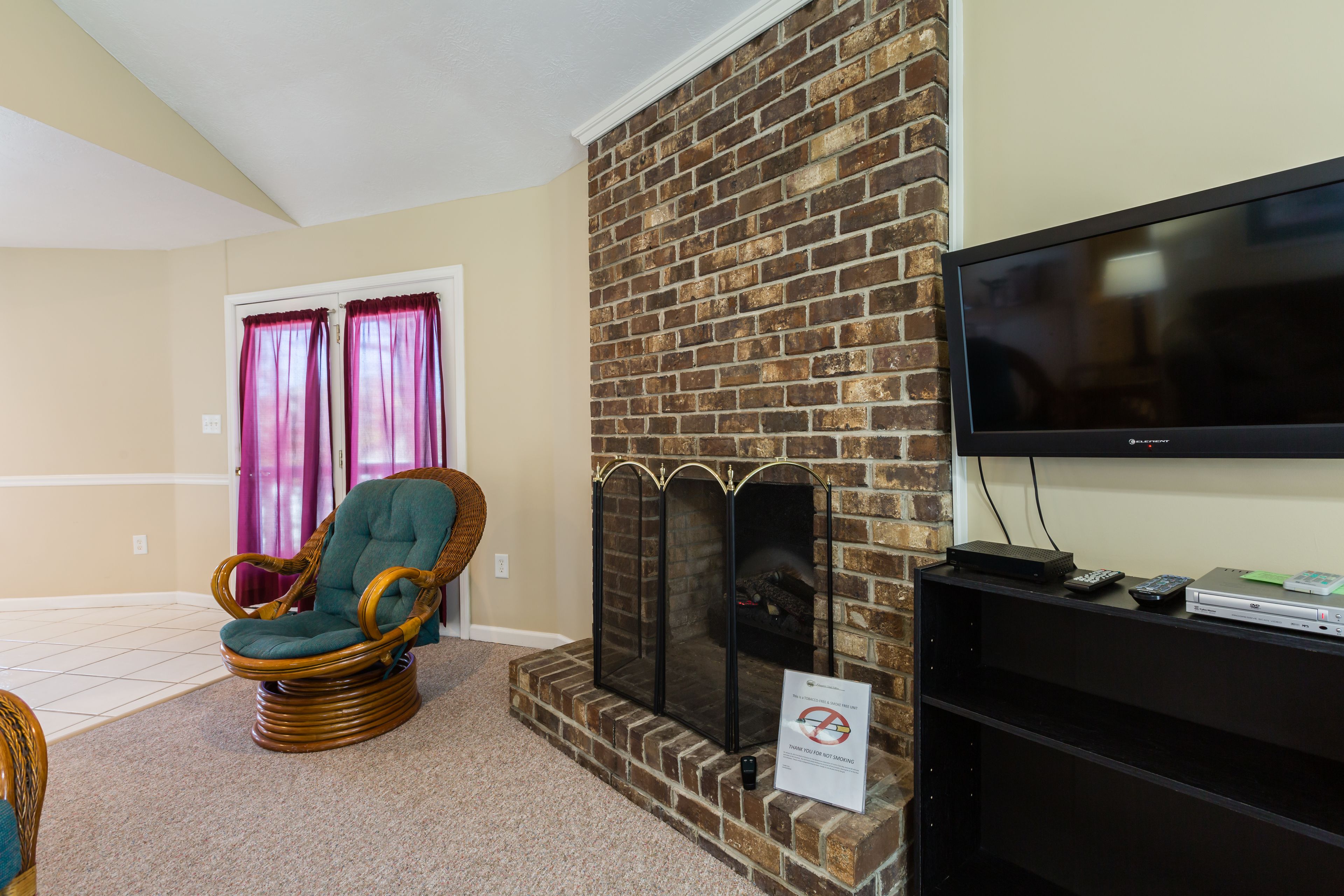 A living room with a brick fireplace, a television mounted on the wall, and a wicker chair.