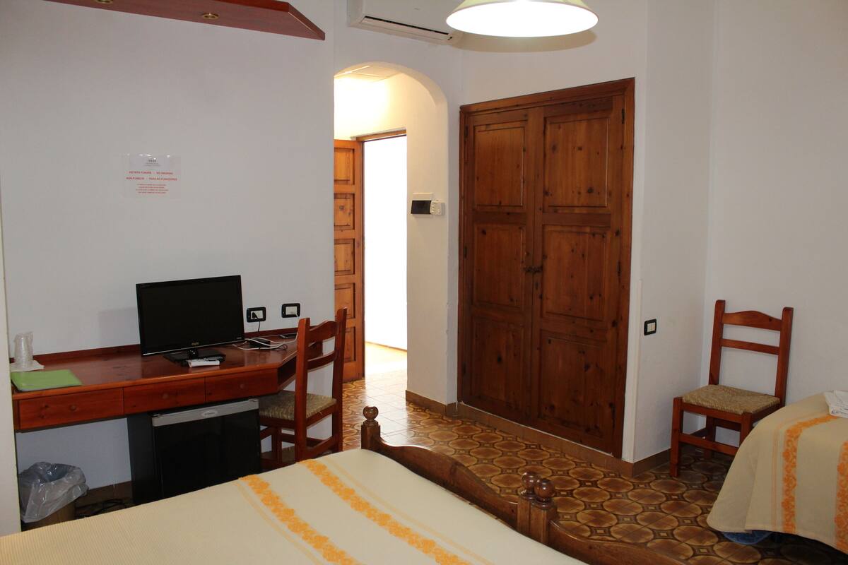 A hotel room with a wooden desk, a television, a bed with a striped bedspread, and a large wooden wardrobe.