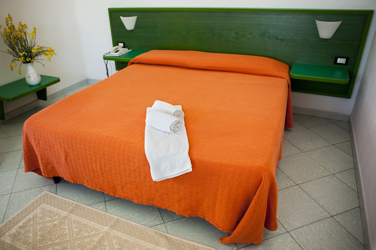 Una cama bien tendida con una colcha naranja y una toalla blanca enrollada, una mesita de noche con un jarrón de flores y un mueble de noche verde con una lámpara.
