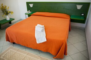 A neatly made bed with an orange bedspread and a rolled white towel, a bedside table with a vase of flowers, and a green nightstand with a lamp. - Hotel Piccada (Palau)