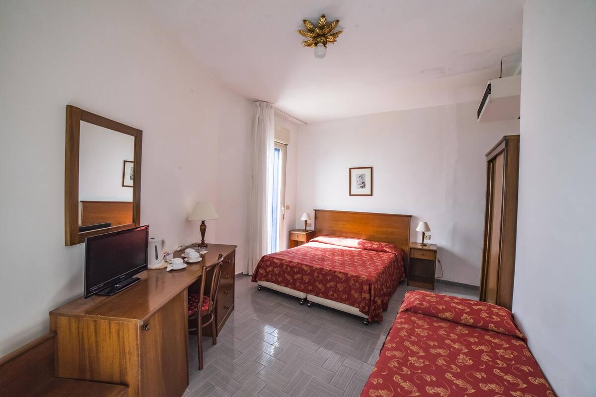 A hotel room with a wooden desk, a television, a bed with red bedding, a bedside table with a lamp, and a framed picture on the wall.
