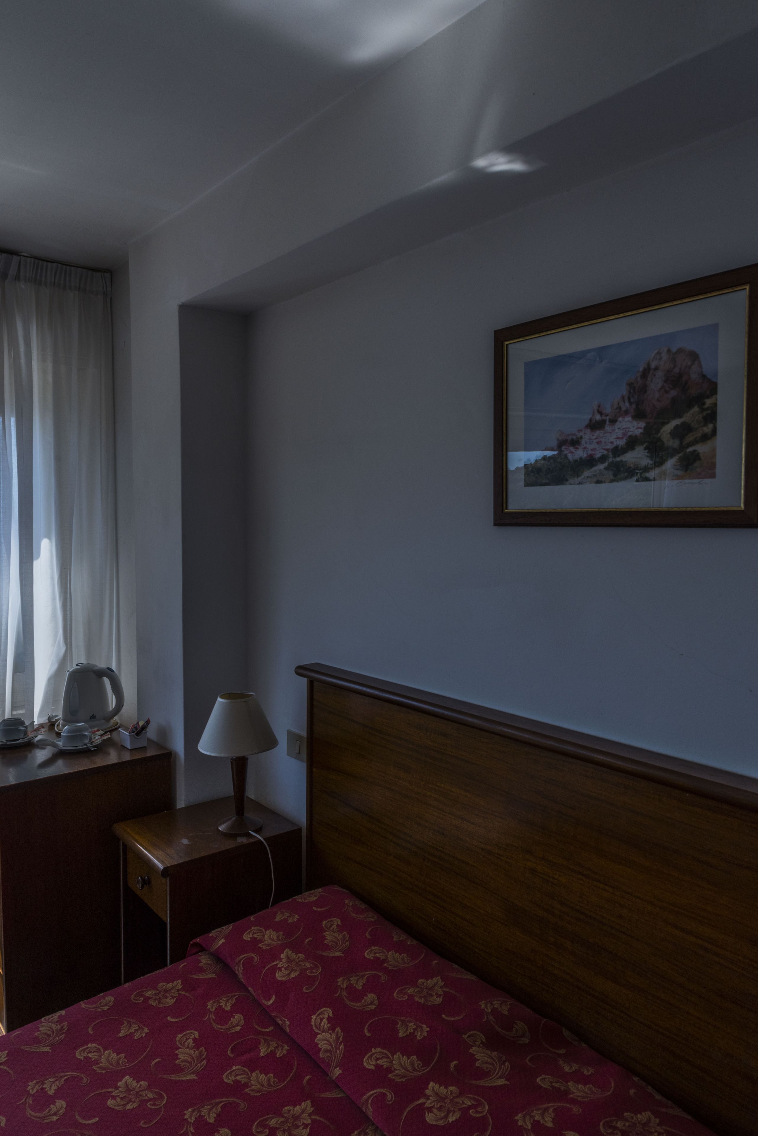 A hotel room with a bed, a nightstand, a lamp, a picture on the wall, and a window with curtains.