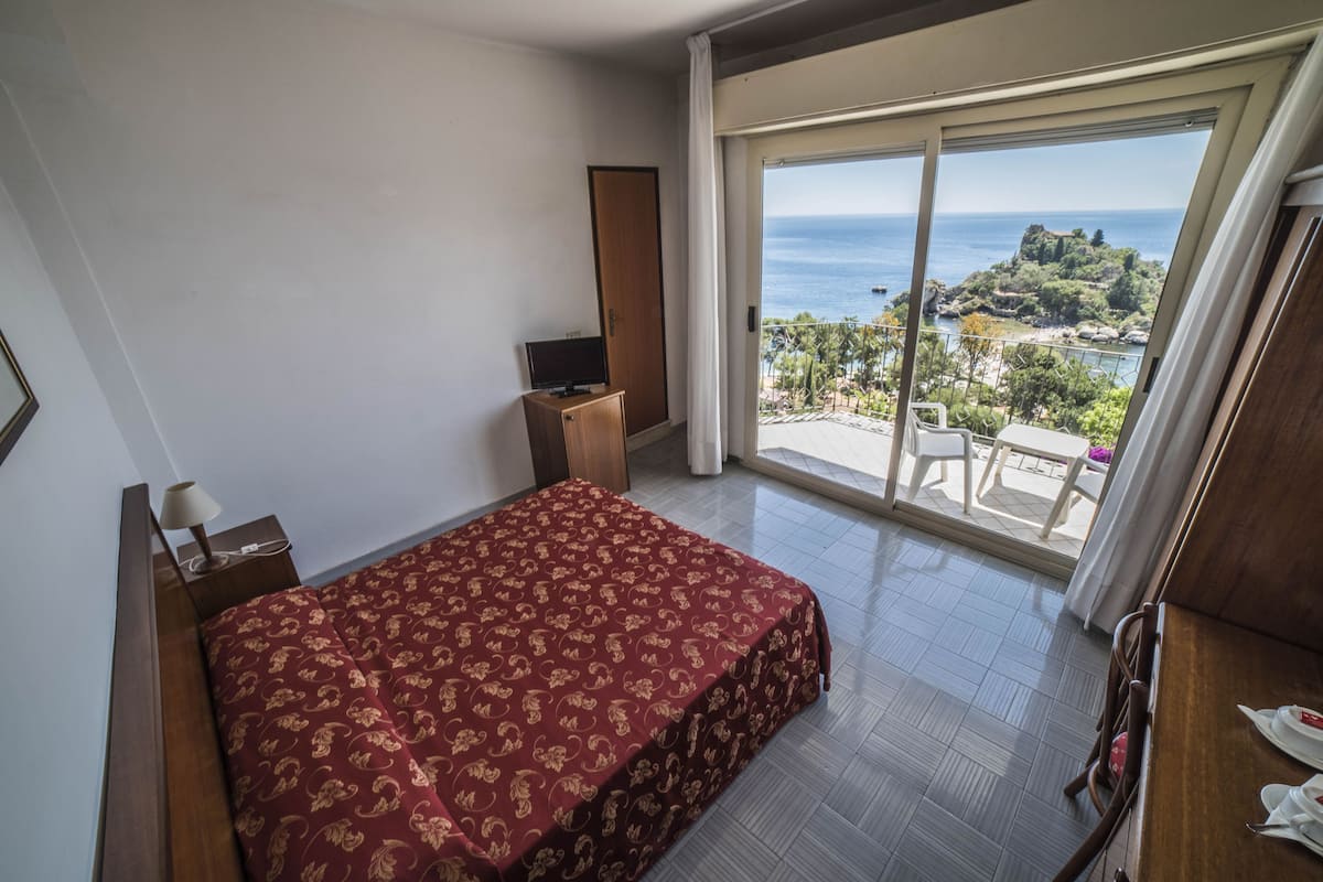 A bedroom with a bed, a TV, and a balcony with a view of the sea.