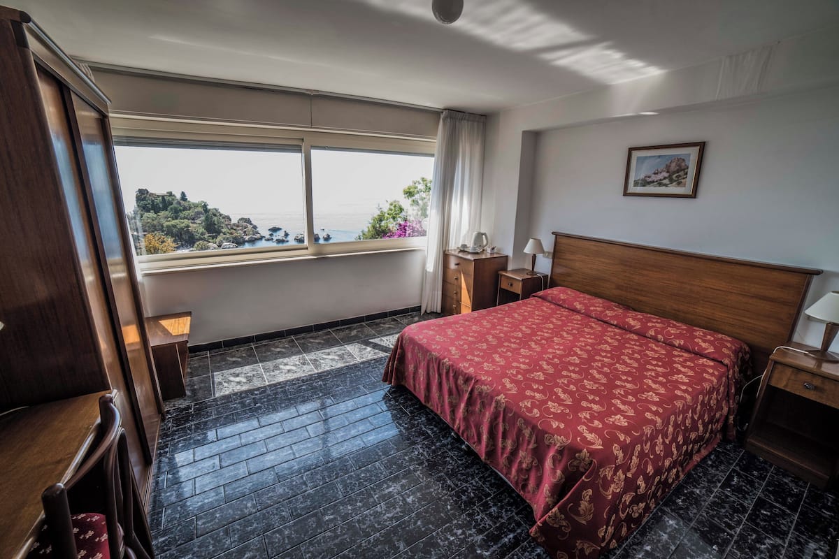 A hotel room with a bed, a nightstand, a lamp, a wooden dresser, and a window with a view of the sea and greenery.