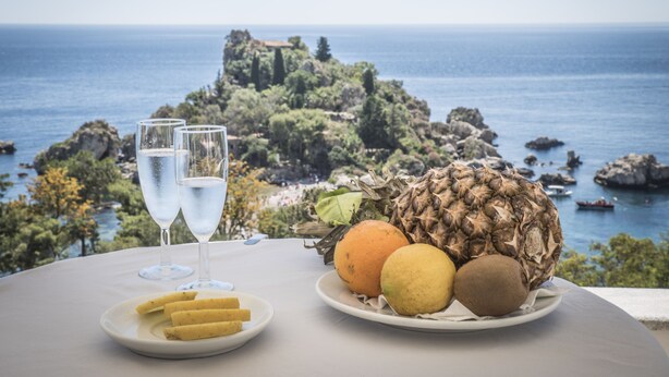 Una mesa con un plato de fruta y dos vasos de agua, con vista a un paisaje costero con afloramientos rocosos y el mar.