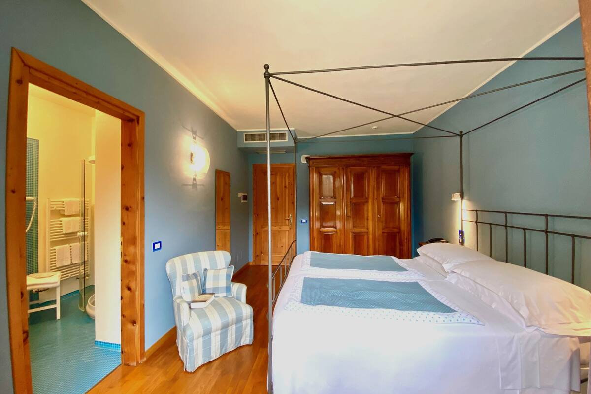 A bedroom with a four-poster bed, a wooden wardrobe, a striped armchair, and a bathroom visible through an open door.