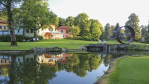 A serene lakeside setting with a reflective pond, a distinctive metal sculpture, and a traditional building with a red roof.