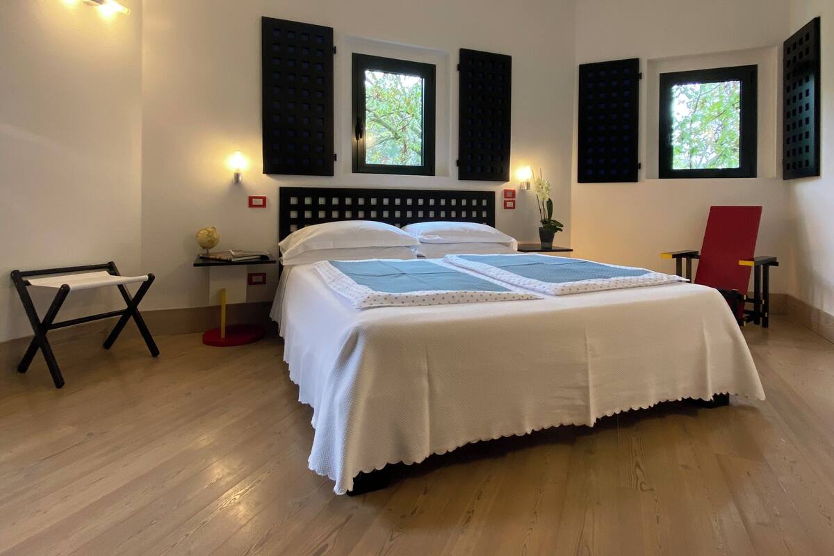 A bedroom with a large bed, two windows with shutters, a bedside table with a lamp, and a small chair.
