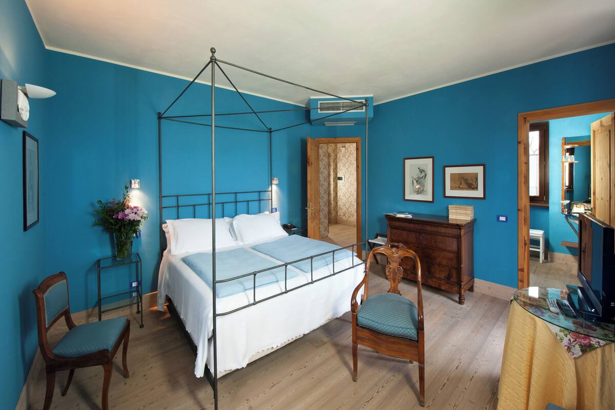 A bedroom with a four-poster bed, a wooden dresser, a chair, a small table, and a mirror.