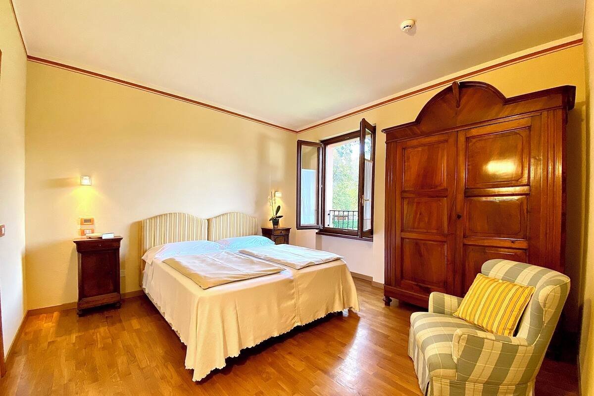 A bedroom with a large bed, a wooden wardrobe, a chair, and a nightstand with a lamp.