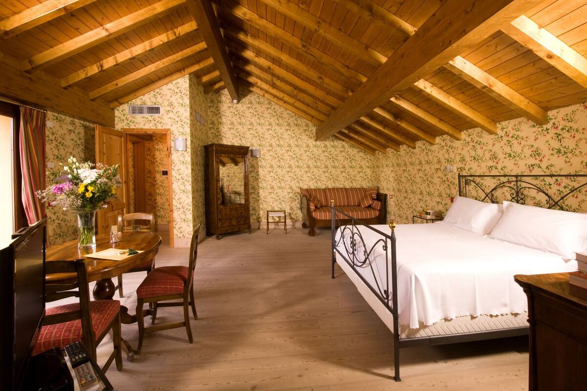 A bedroom with a wooden ceiling, a bed, a nightstand, a chair, a table with a vase of flowers, and a wooden dresser.