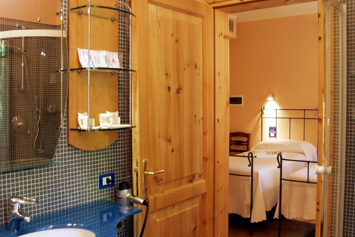 A bathroom with a blue sink, a glass shower enclosure, and a wooden door leading to a bedroom with a bed and a nightstand.