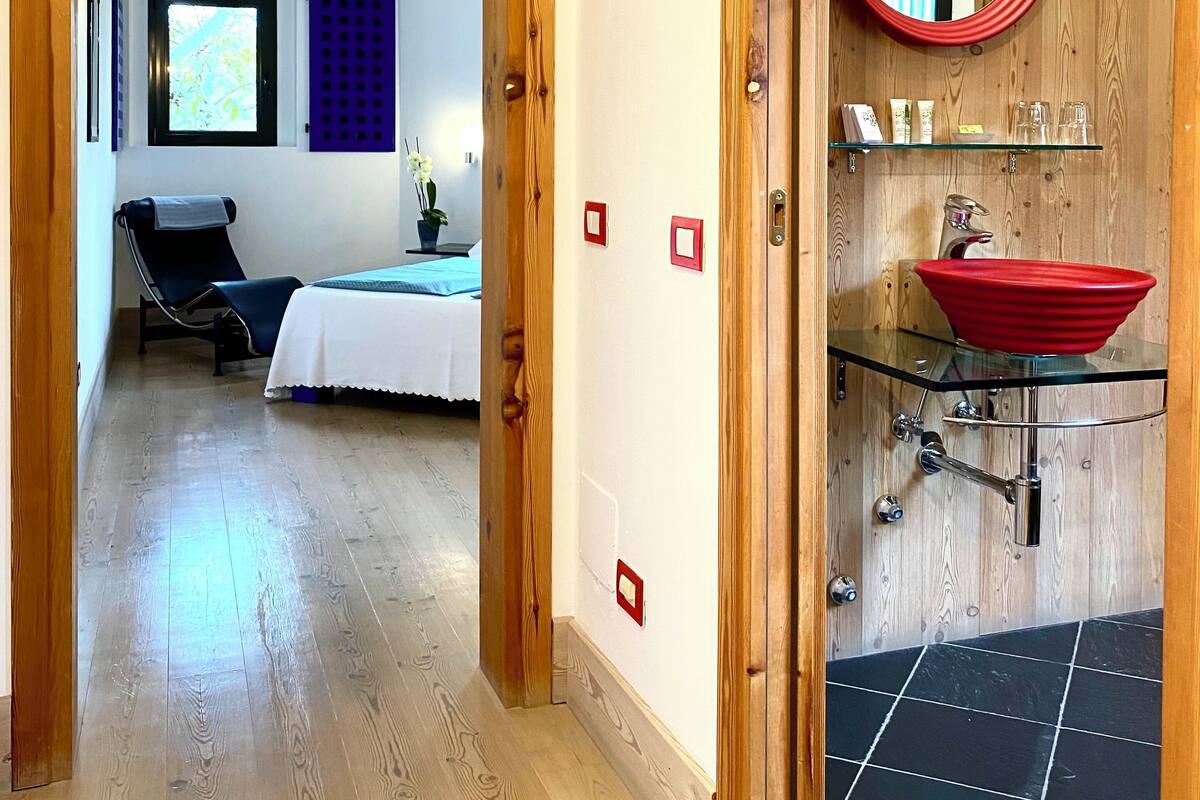 A hotel room with a wooden floor, a bed with a white bedspread, a chair, a window with a view, and a bathroom with a red sink and mirror.