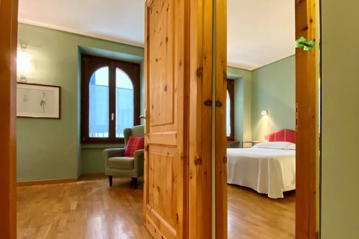 A room with wooden flooring, a bed with white bedding, a green armchair, and a window with a view.