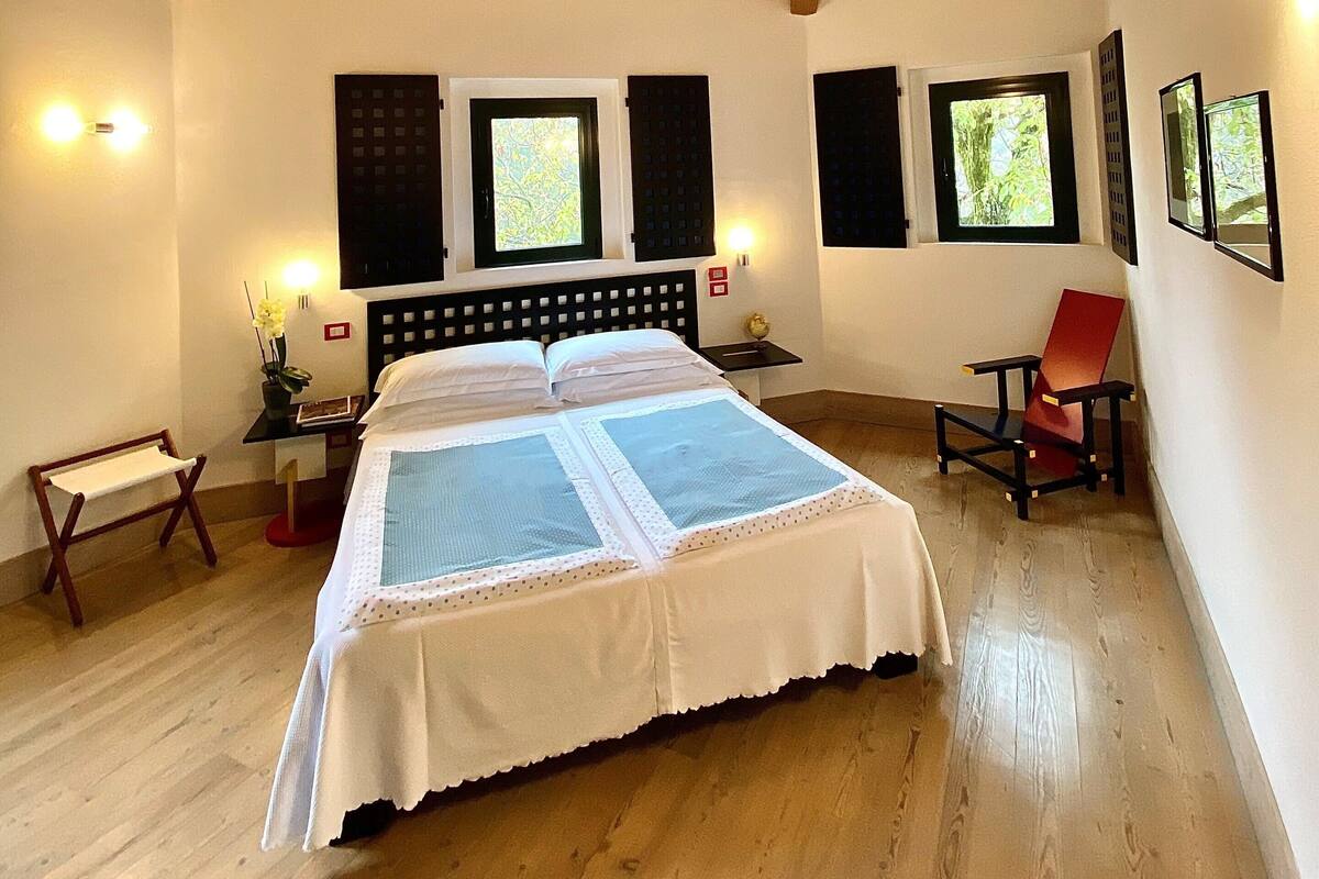 A bedroom with a wooden ceiling, a large bed, two windows, a chair, and a bedside table.