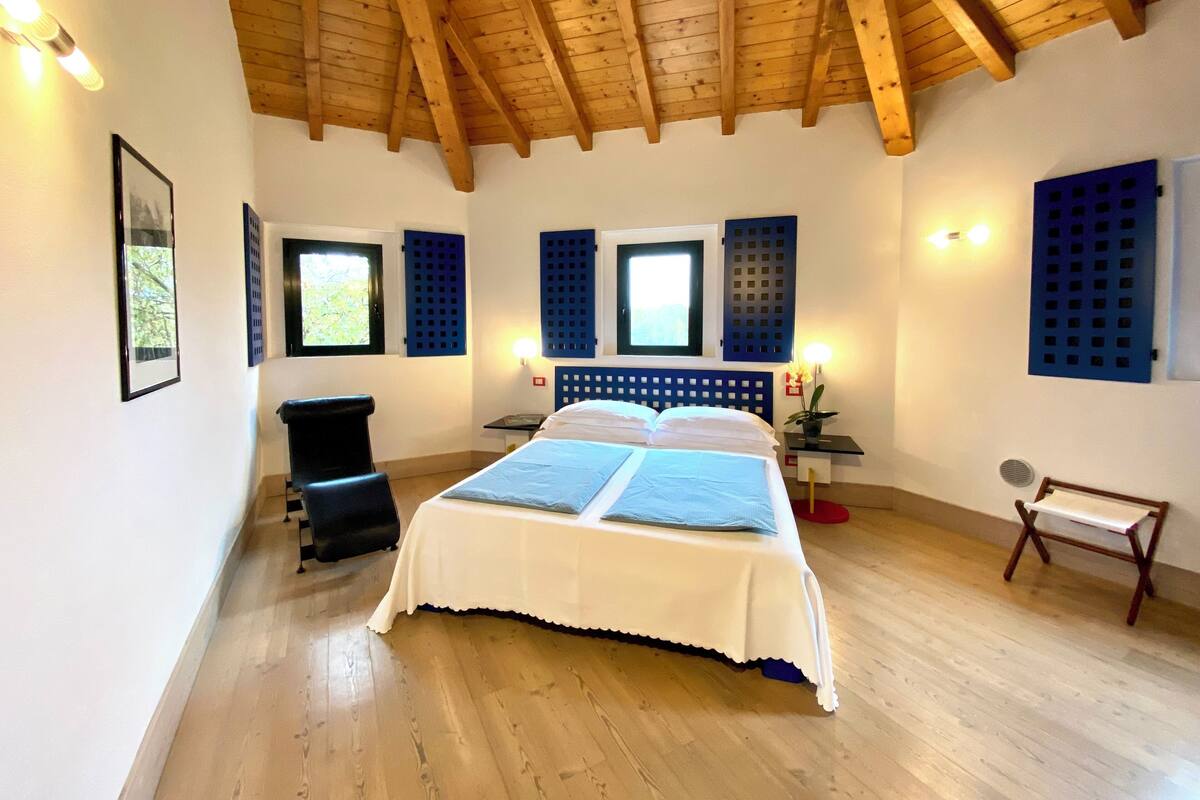 A bedroom with a wooden ceiling, a bed with white bedding, two windows with blue shutters, a black chair, and a small table with a red lamp.