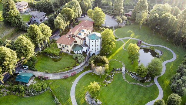 An aerial view of a large estate with a central building, multiple guest rooms, and a well-maintained garden with a pond and winding paths.