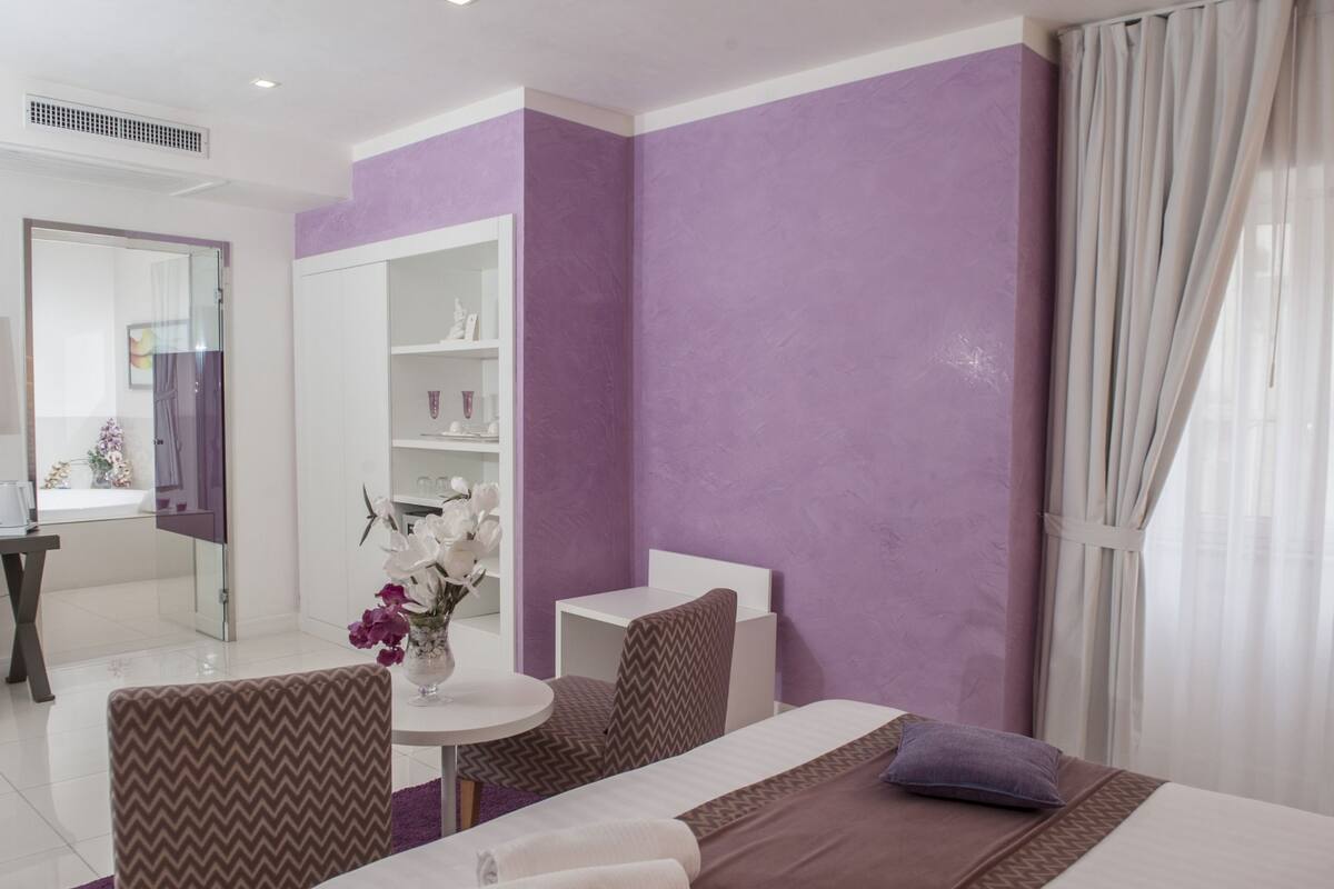 A hotel room with two beds, a small round table, a chair, a vanity with a mirror, and a bathroom visible through an open door.