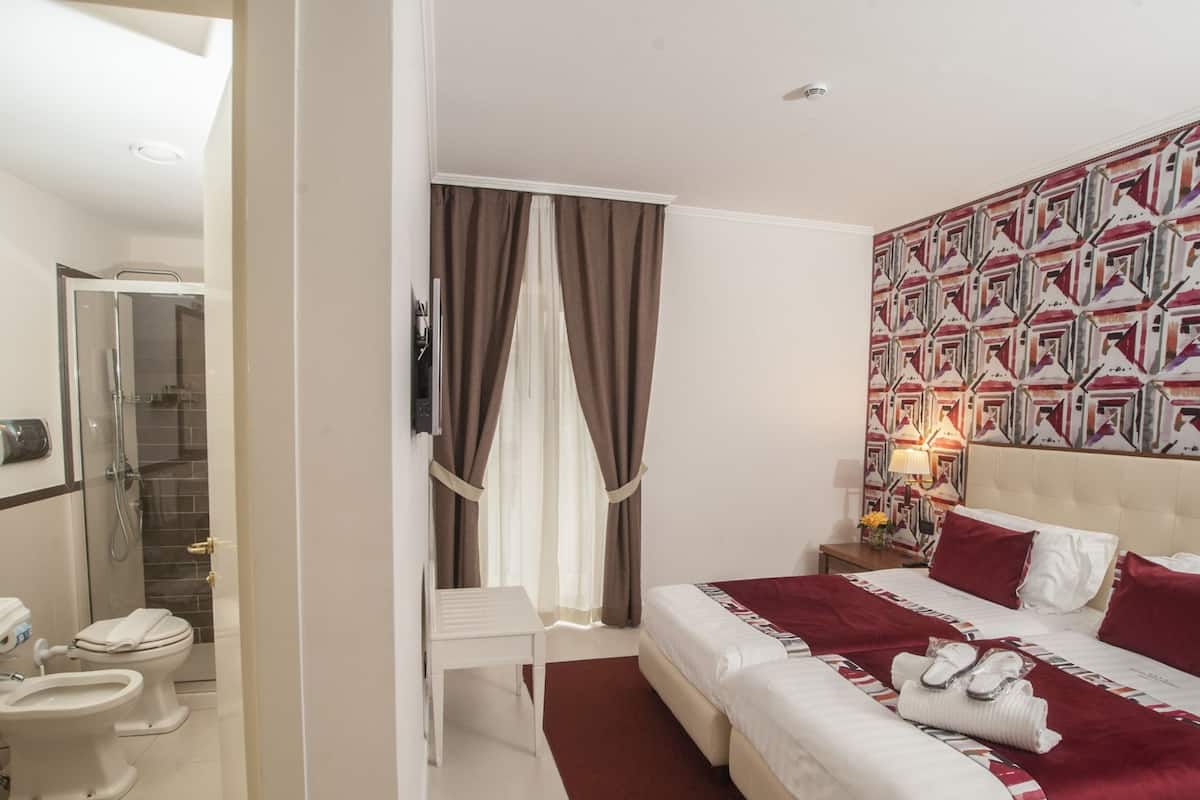 A hotel room with a bed, a bedside table with a lamp, a shower area, and a bathroom with a toilet and sink.