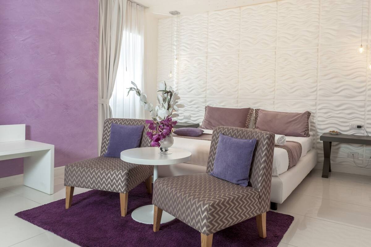 A modern living room with a white sofa, two patterned armchairs, a round coffee table, and a purple rug.