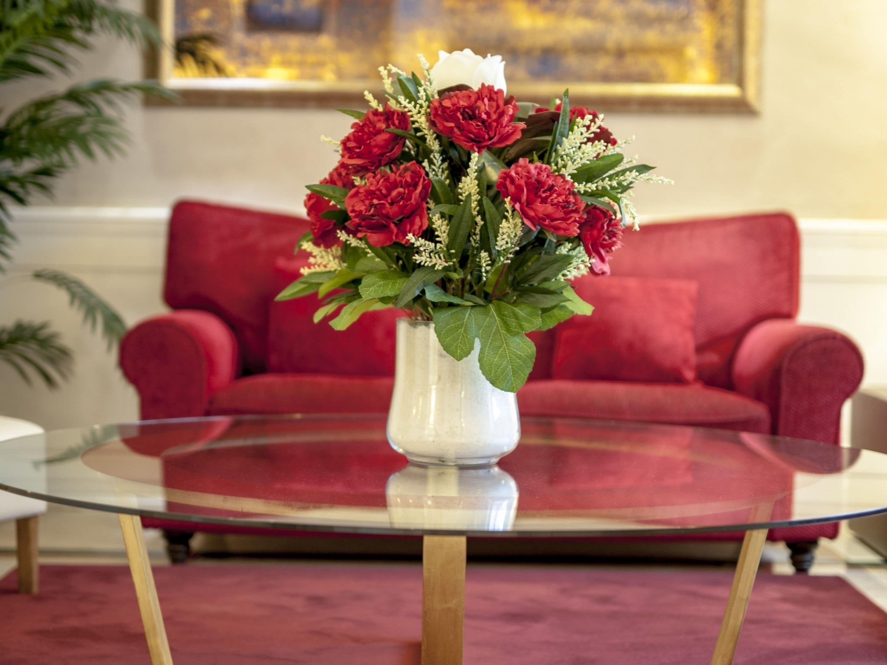 A round glass table with a white vase containing red and white flowers, positioned in front of a red sofa.