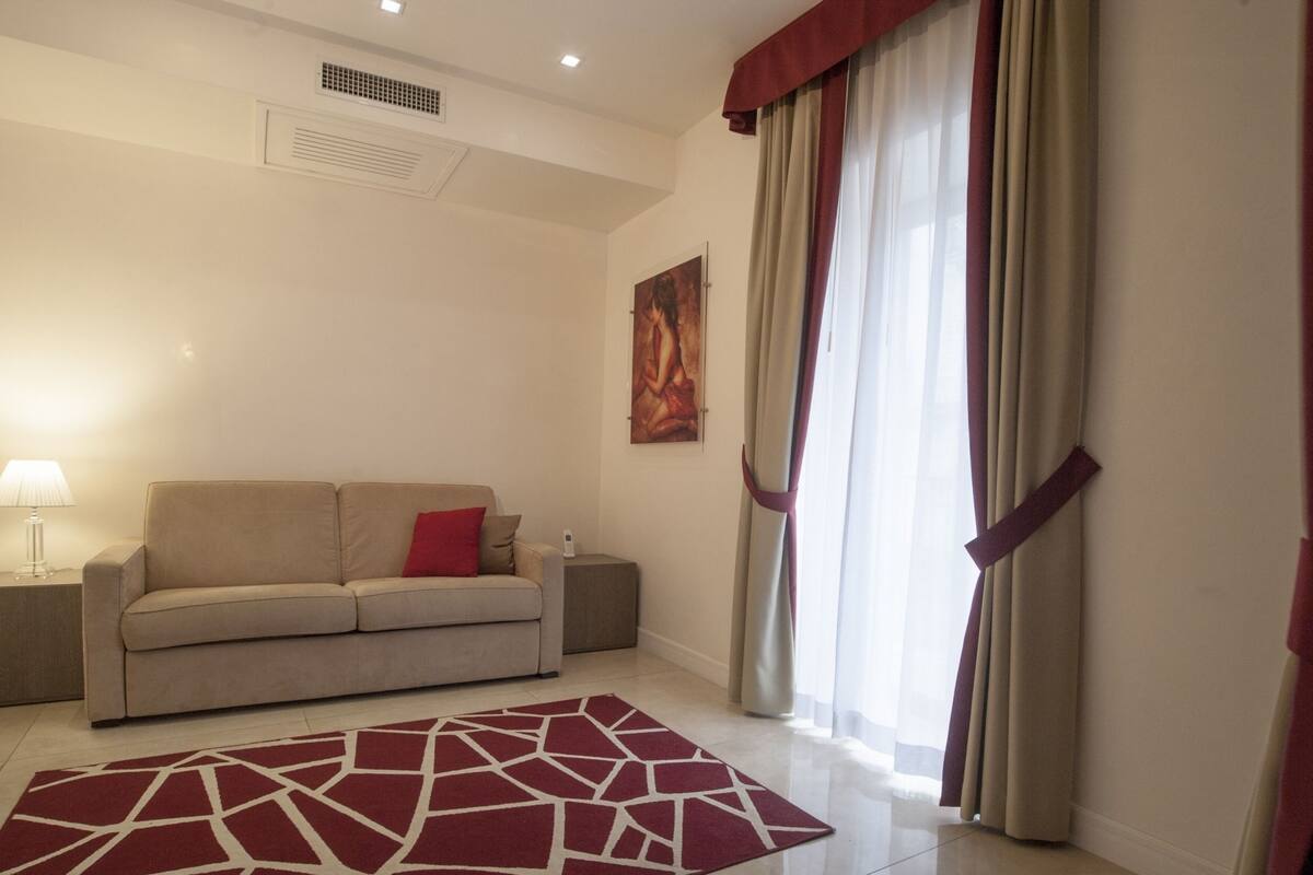 A living room with a beige sofa, a red and white rug, a painting on the wall, and a window with curtains.