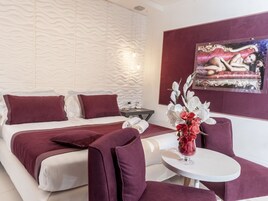 Habitación de hotel moderna con una cama grande, una mesita pequeña y una silla. Hay un cuadro en la pared y un florero con flores sobre la mesita.