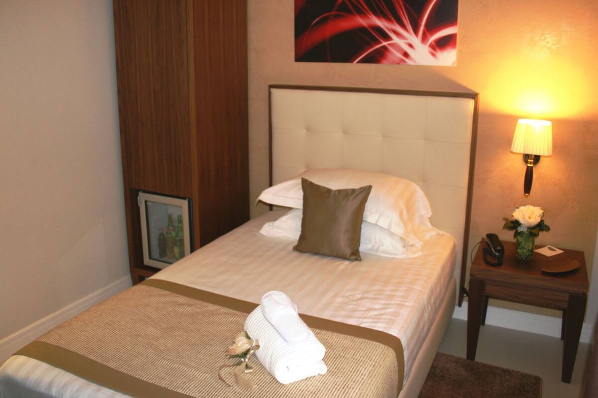 A hotel room with a neatly made bed, a bedside lamp, a small table with a vase of flowers, and a framed picture on the nightstand.