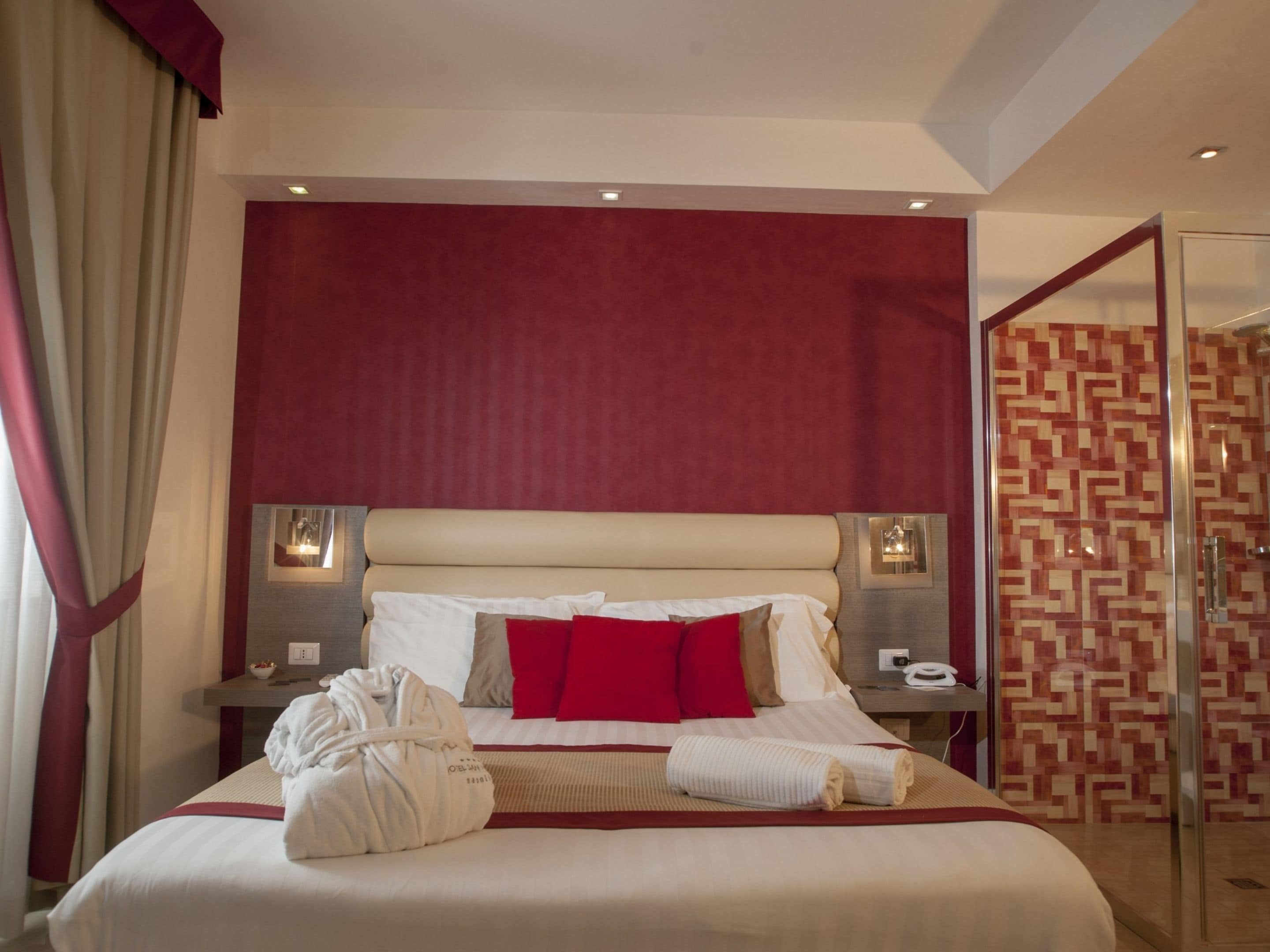 A hotel room with a large bed, red curtains, a red wall, a glass shower, and a patterned tile wall.