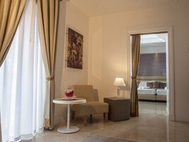 Una camera d'albergo con angolo salotto, un tavolino con una lampada, un quadro appeso al muro e vista su un'altra stanza con un letto.
