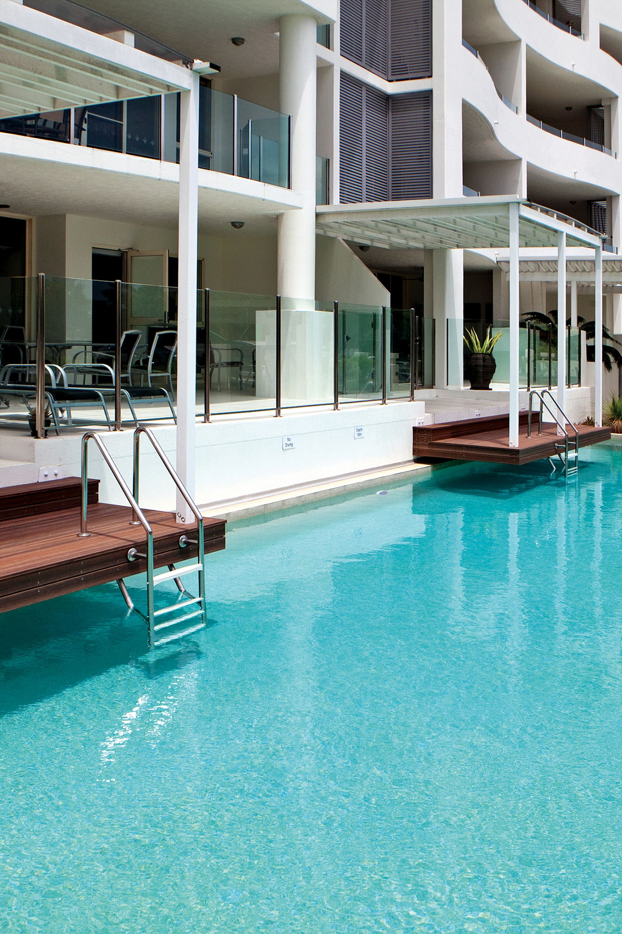 Une piscine dotée d’une terrasse en bois et de rampes en métal, entourée d’un bâtiment moderne avec des balcons et des garde-corps en verre.