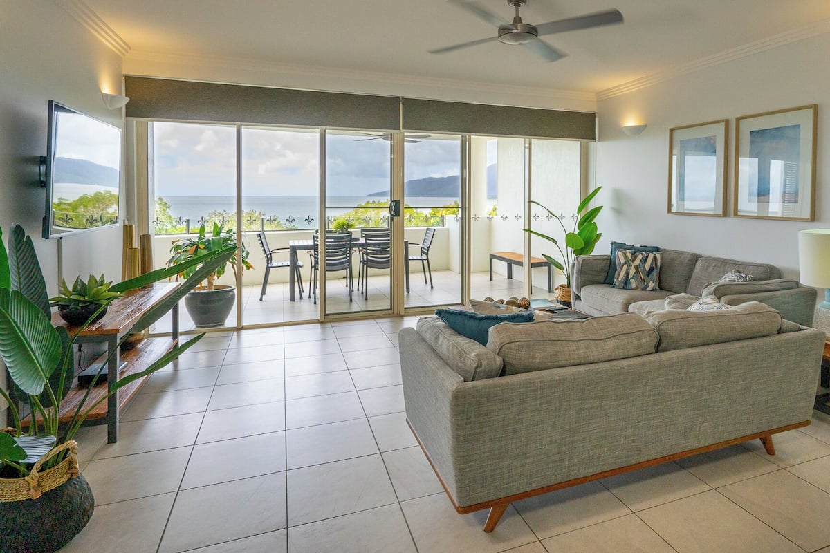 A spacious living area with a large sliding glass door leading to an outdoor balcony, a grey sofa, a small table with a lamp, and several potted plants.