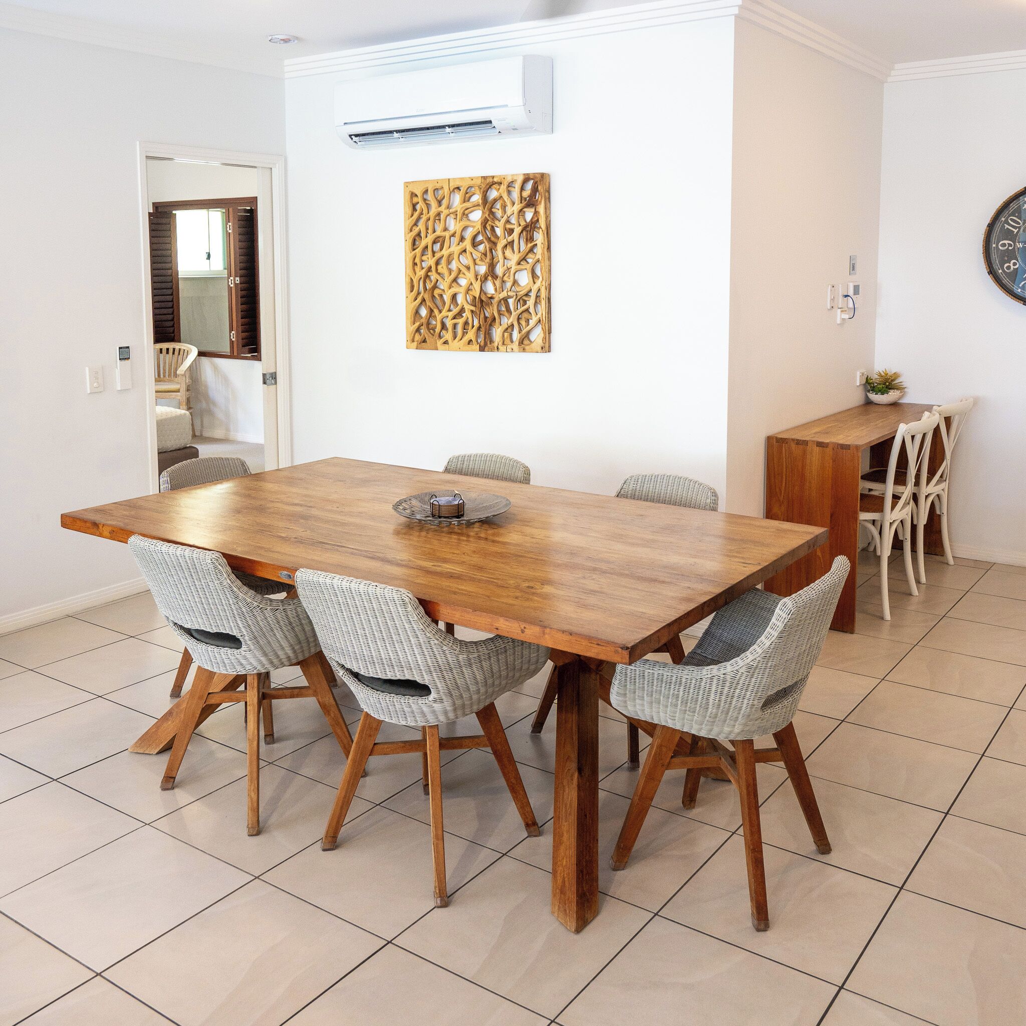 Een eetgedeelte met een houten tafel, rieten stoelen, een decoratief kunstwerk en een airconditioning.