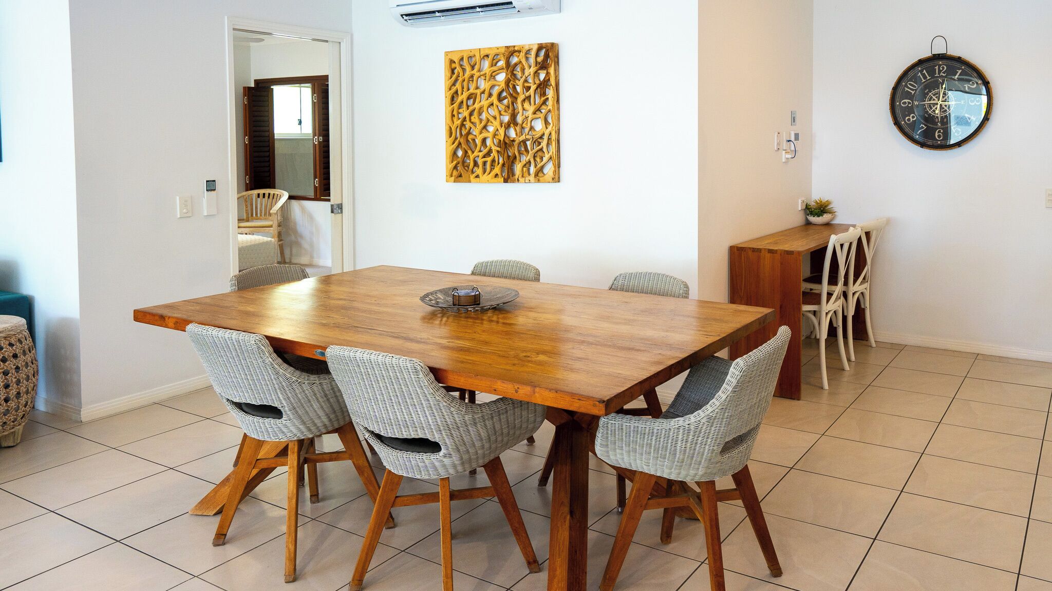A dining area with a wooden table and wicker chairs, a decorative wall piece, and a wall clock.