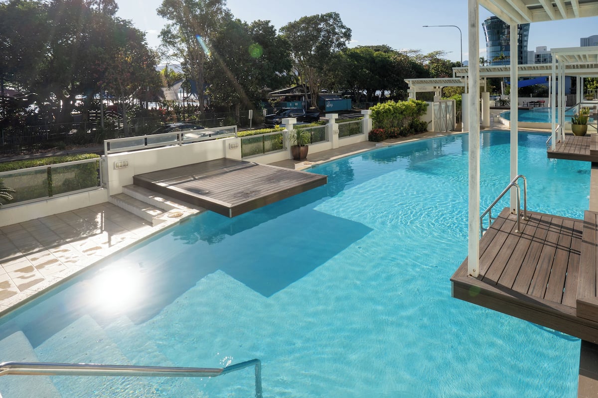 A clear blue pool with wooden deck areas, surrounded by modern buildings and lush greenery.