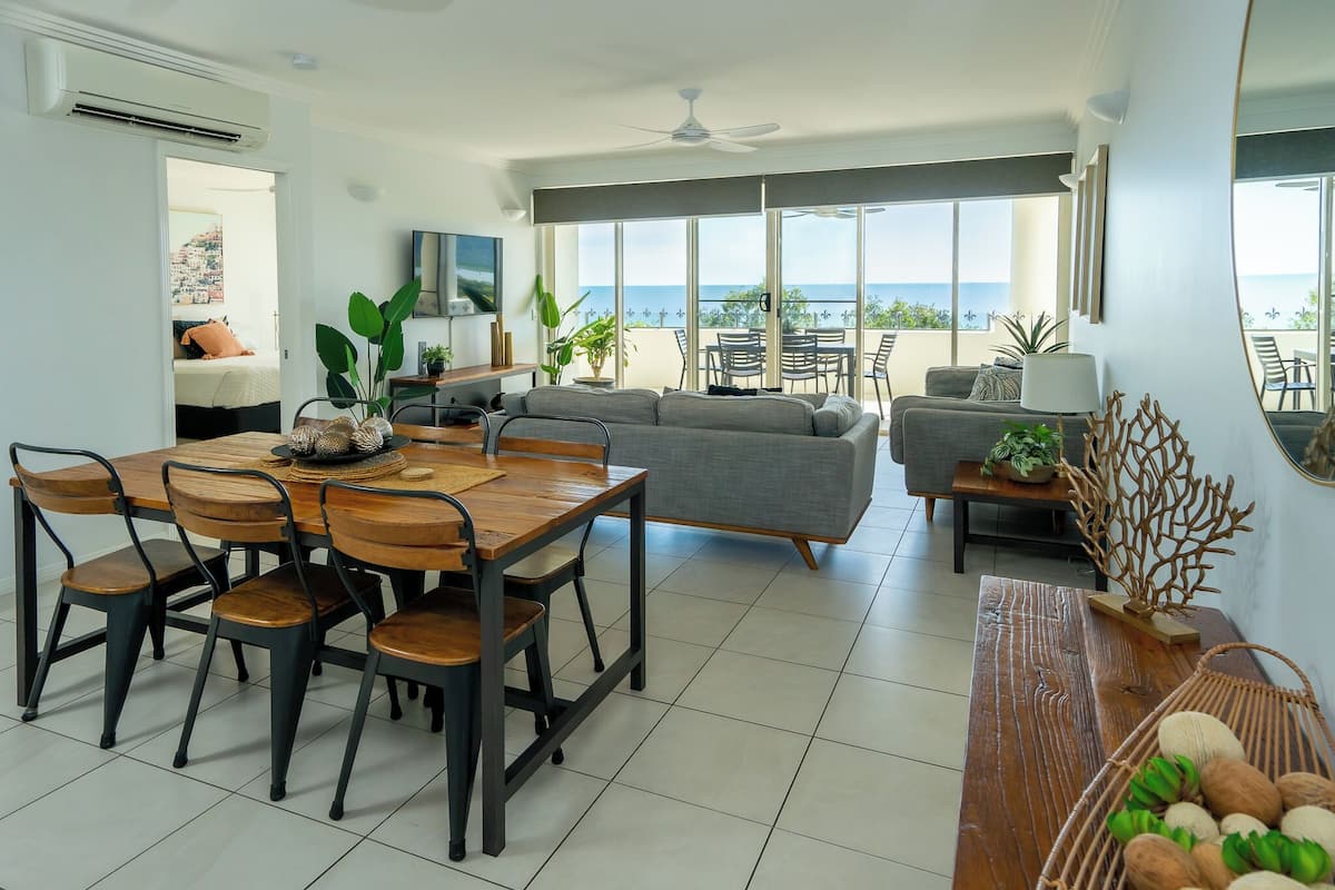 A spacious living area with a dining table, sofa, and chairs. A large window offers a view of the ocean.