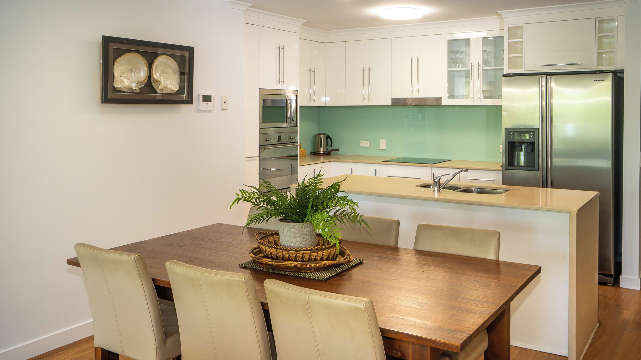 Una cocina moderna con armarios blancos, un refrigerador de acero inoxidable, un microondas y un lavamanos. Hay una mesa de comedor con sillas y una planta en maceta sobre la mesa.