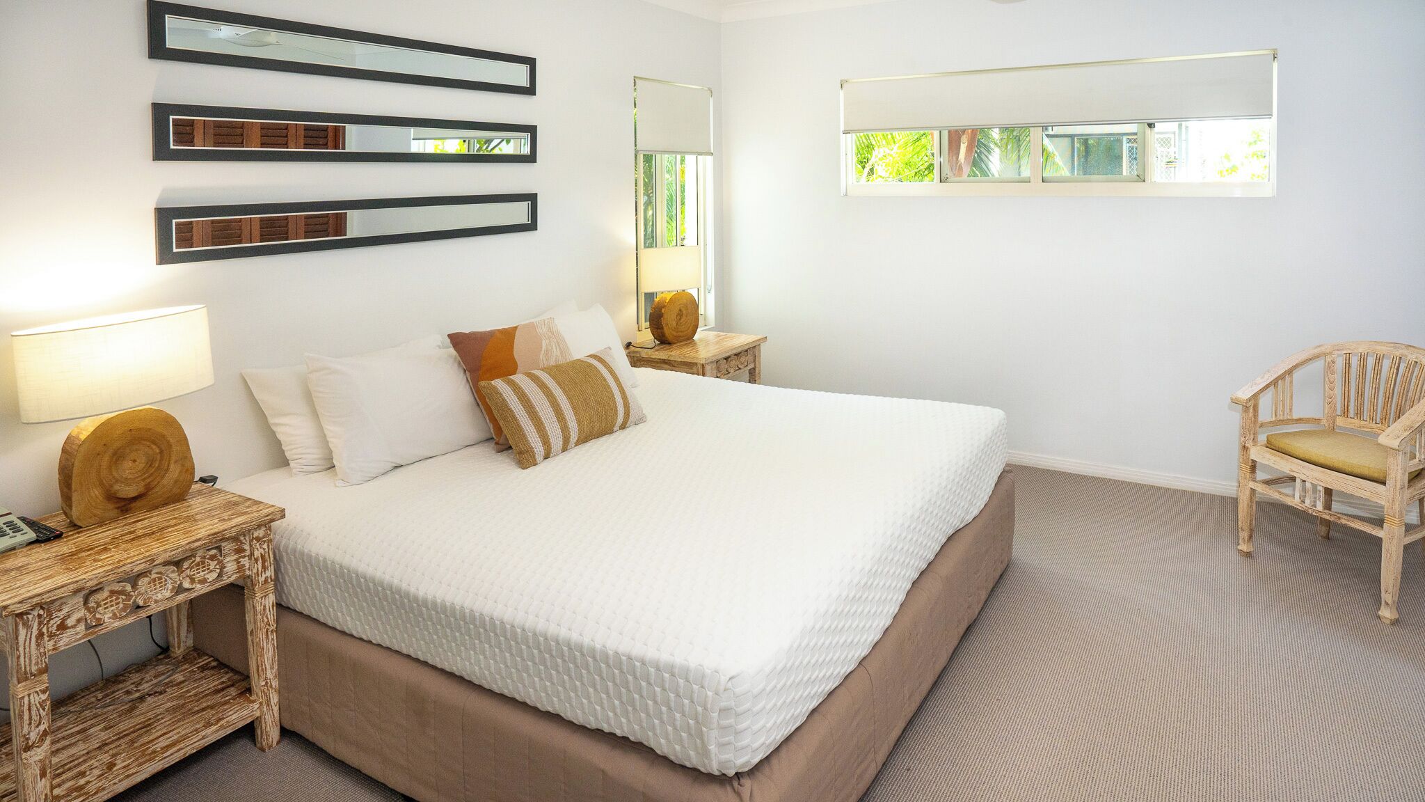 A neatly arranged bedroom with a large bed, a bedside table with a lamp, a chair, and three wall-mounted mirrors.
