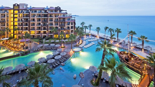 Un resort con varias piscinas, palmeras y un edificio grande con balcones con vista al océano.