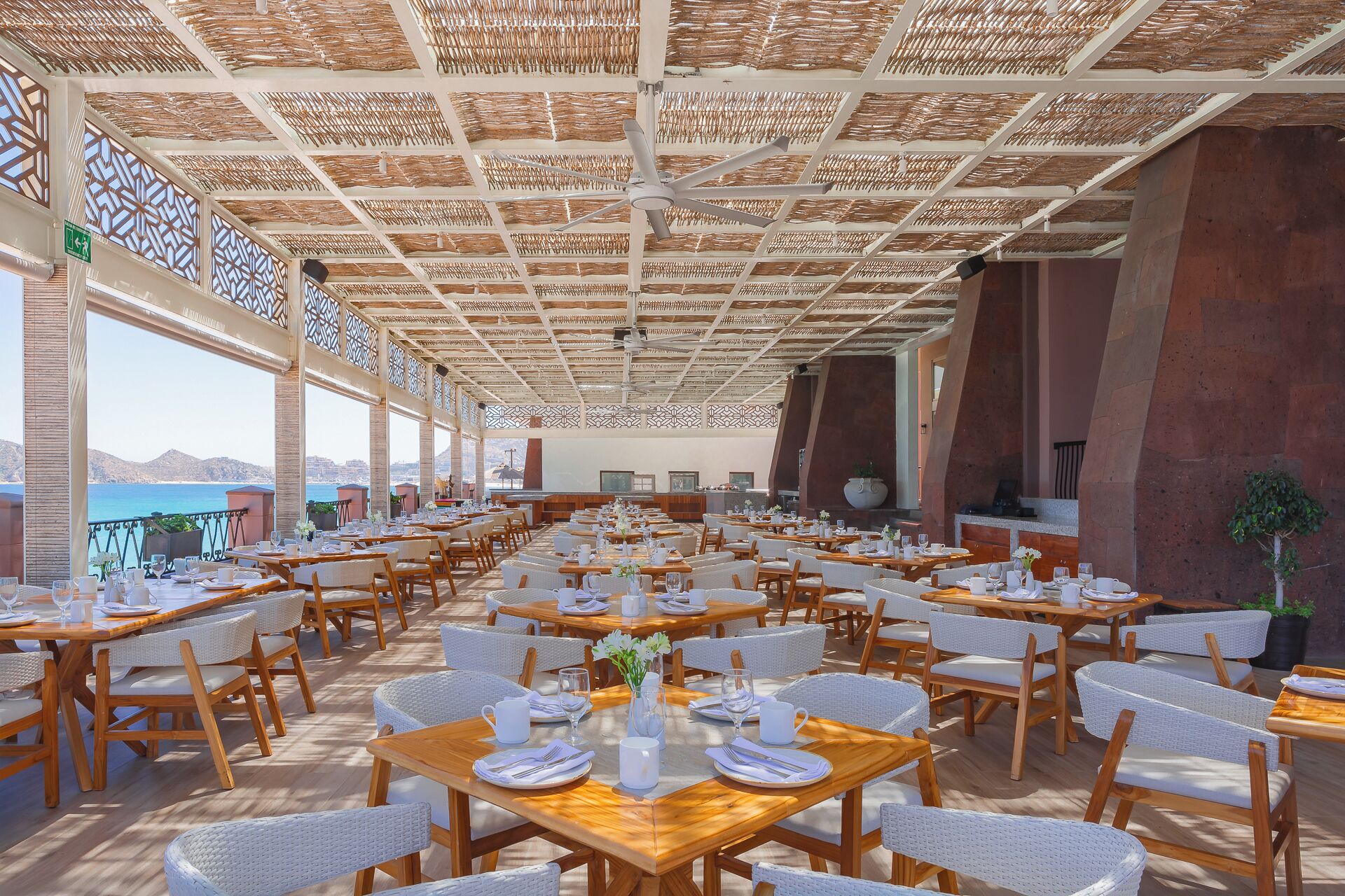 Un amplio espacio de comedor al aire libre con varias mesas y sillas, techo con diseño tejido y vista al mar.