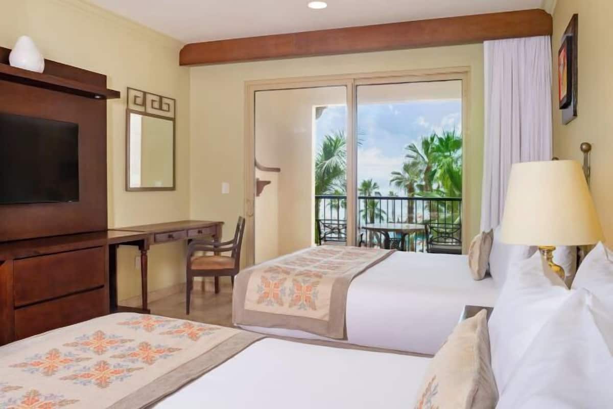 Habitación de hotel con dos camas, un escritorio, una silla, un televisor, un balcón con vistas a palmeras y una ventana con cortinas.