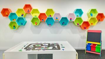 Ein Zimmer mit einem weißen Tisch, einer Spielunterlage mit Stadtmotiv, bunten sechseckigen Regalen mit Spielzeug und einer Spielzeugaufbewahrungsbox mit schwarzer Oberseite.