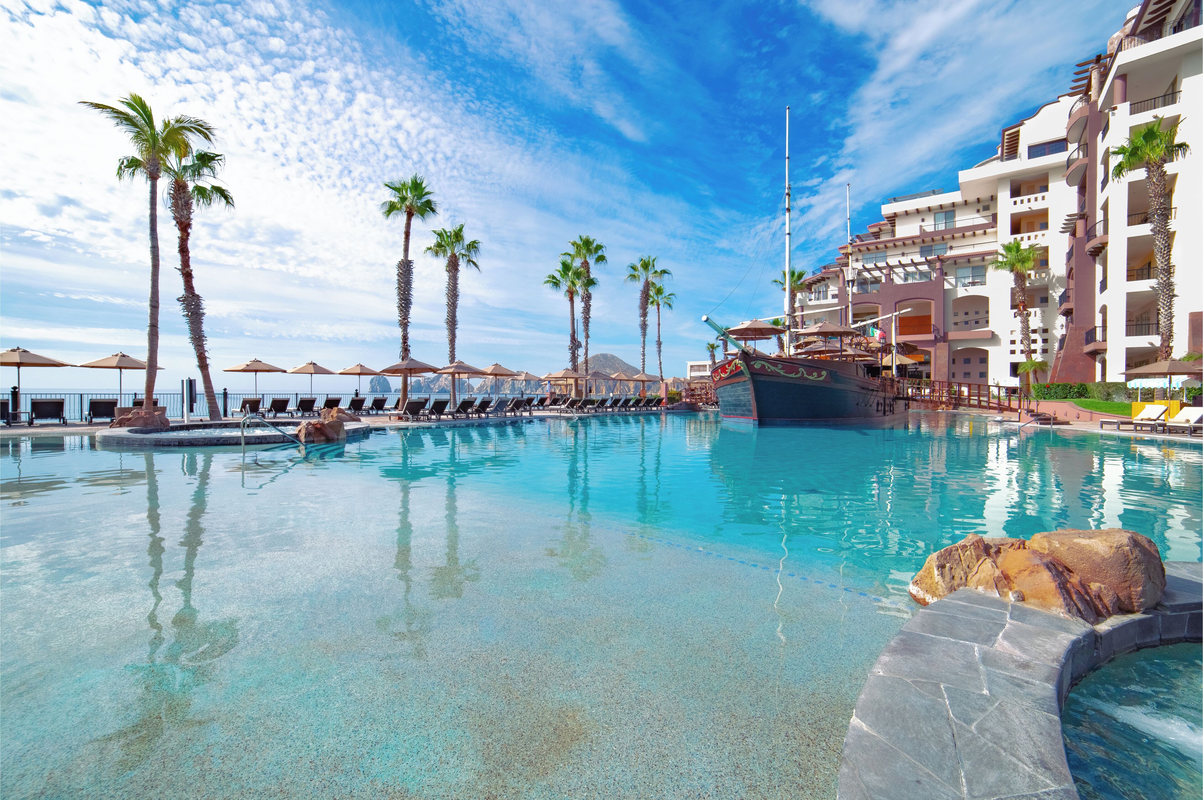 Une piscine d’hôtel avec des palmiers, des chaises longues et un bateau.