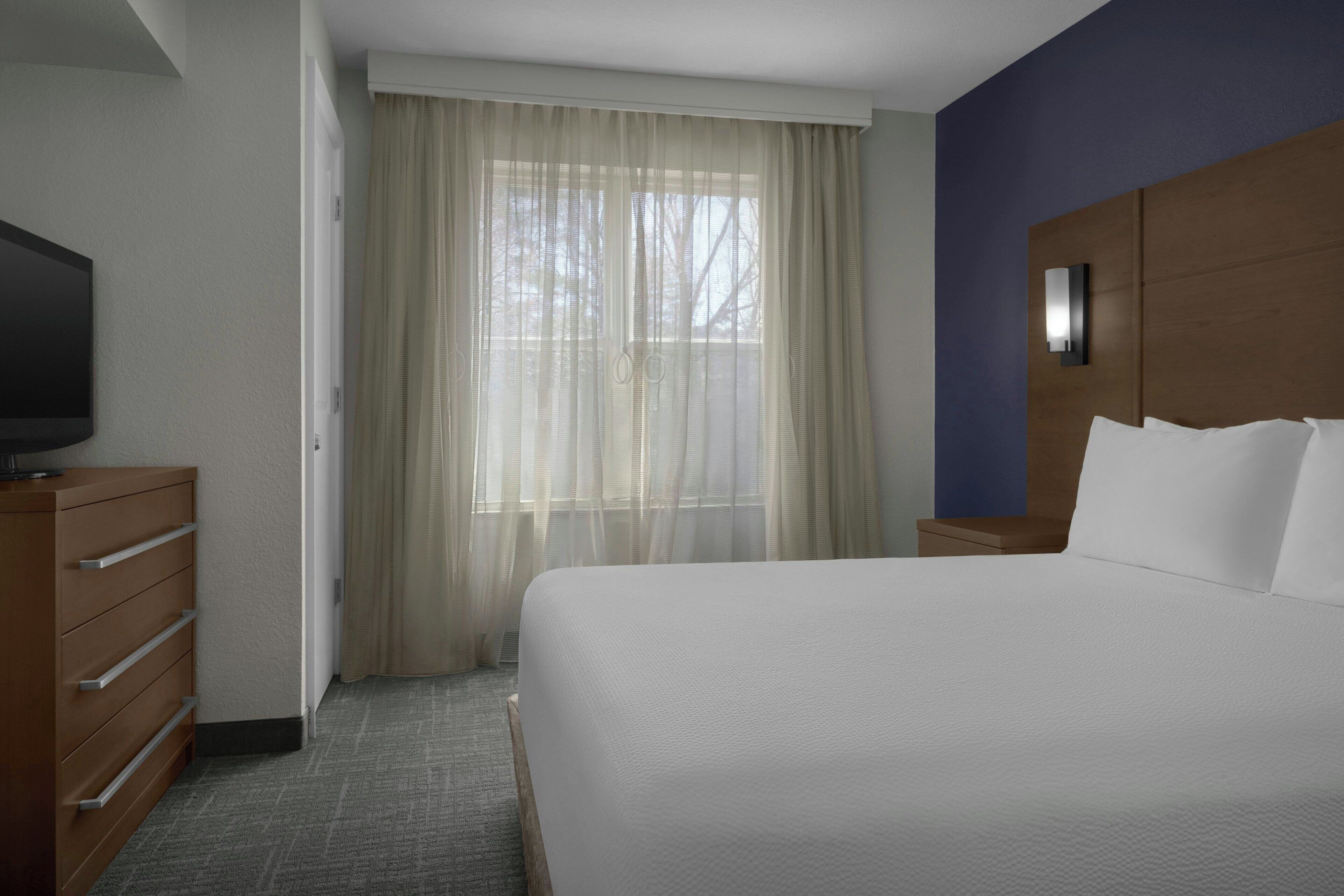 A hotel room with a bed, a television on a dresser, a window with sheer curtains, and a nightstand with a lamp.