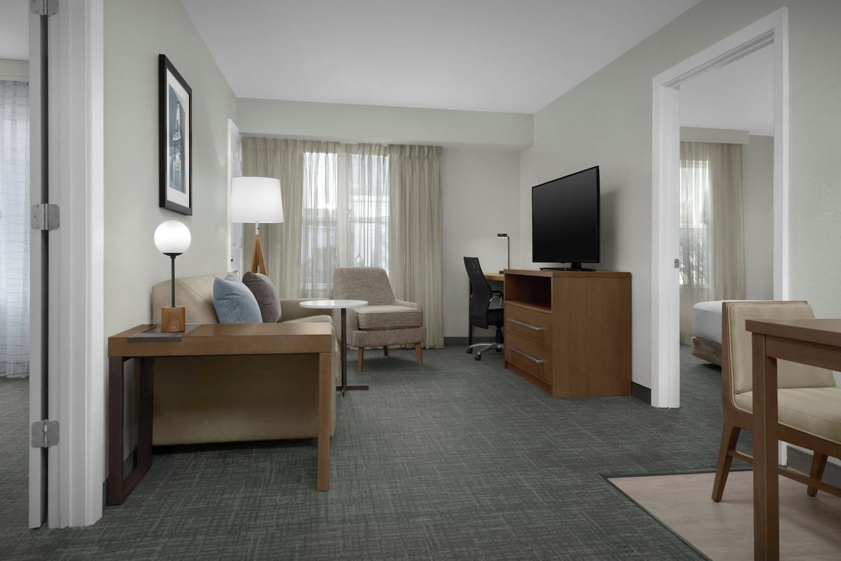A hotel room with a sofa, armchair, desk, television, and a view of the outdoors.