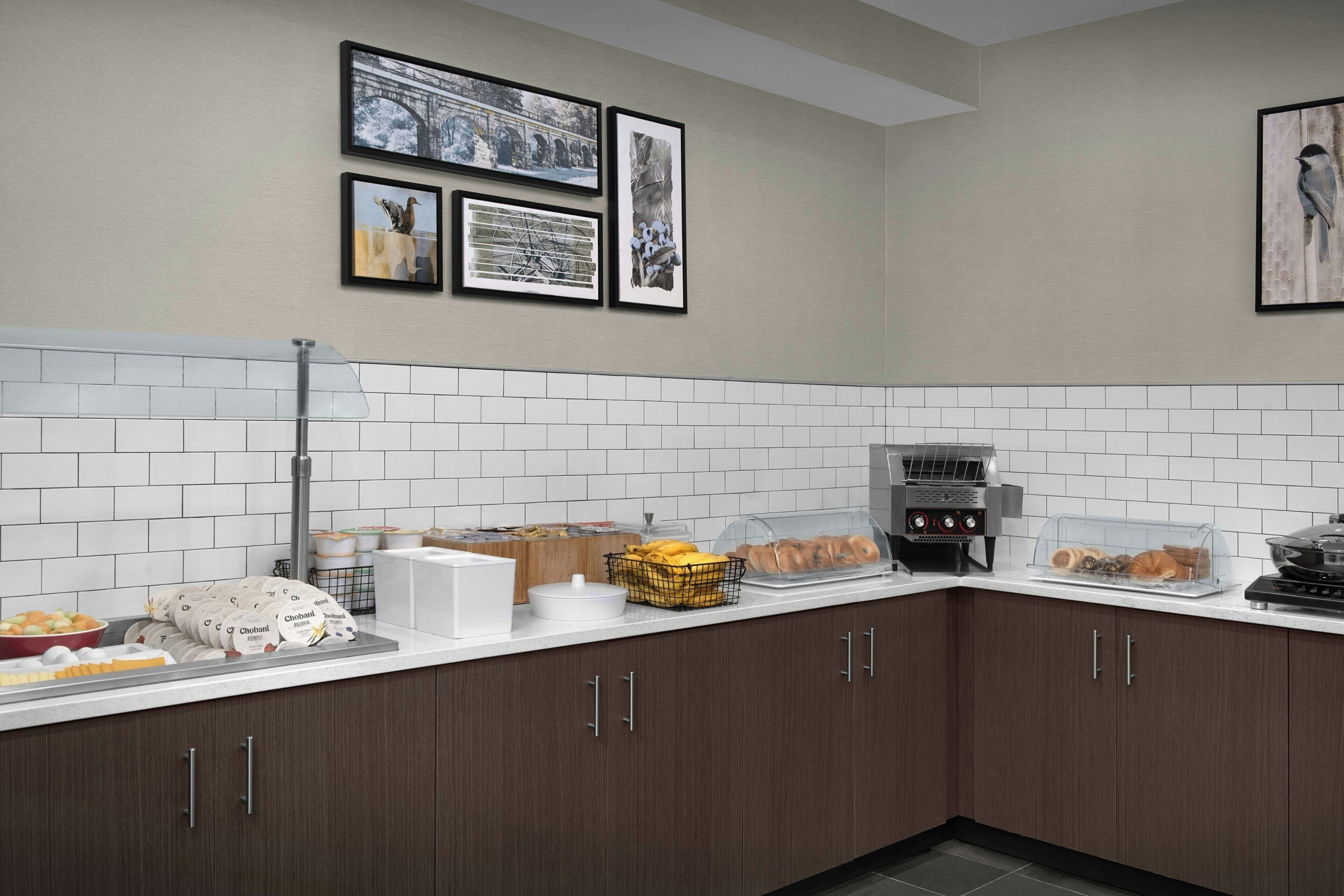 Uma cozinha com painel de azulejos brancos, armários de madeira escura, uma cafeteira e diversos alimentos sobre o balcão.