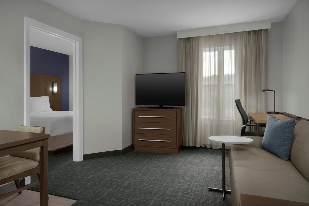 A hotel room with a bed, a TV on a wooden cabinet, a small table, a sofa, and a window with curtains.