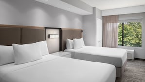 A hotel room with two beds, a nightstand, a window with curtains, and a view of trees.