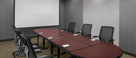 Un salón de conferencias con una mesa larga, seis sillas, una pantalla de proyector e iluminación moderna.