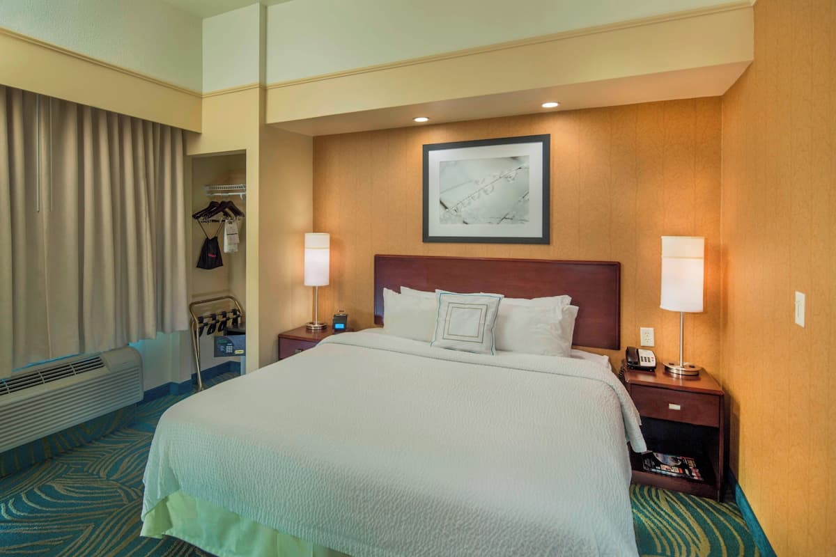 Quarto de hotel com uma cama grande, duas luminárias de cabeceira, uma moldura na parede, um porta-roupas com peças penduradas e uma mesinha com telefone e revista.